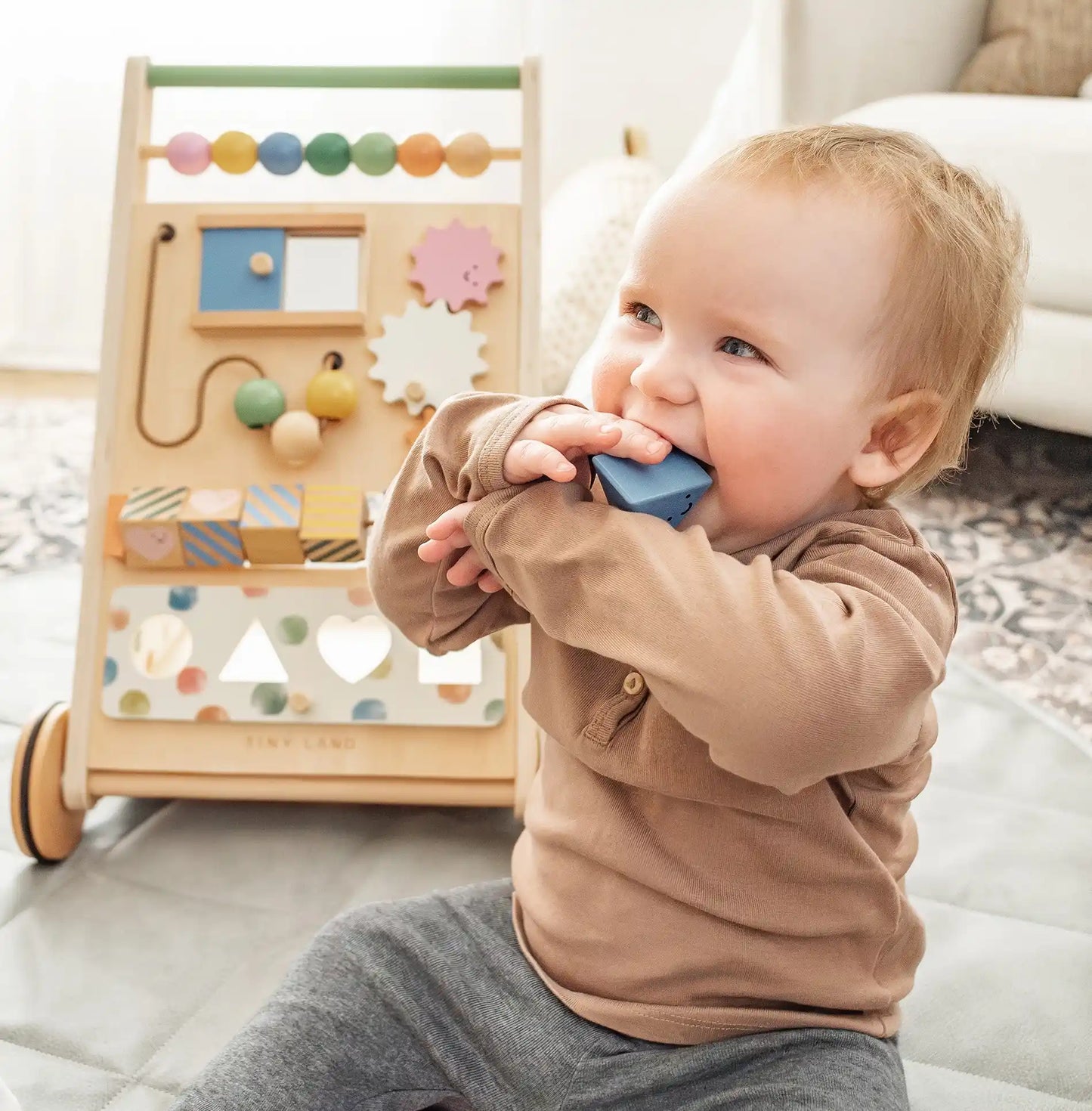 A smiling toddler in a brown shirt and gray pants sits on a mat, chewing a blue block, with the Tiny Land® Versatile Natural Wooden Baby Walker—featuring colorful beads and gears—placed behind them for sensory play.