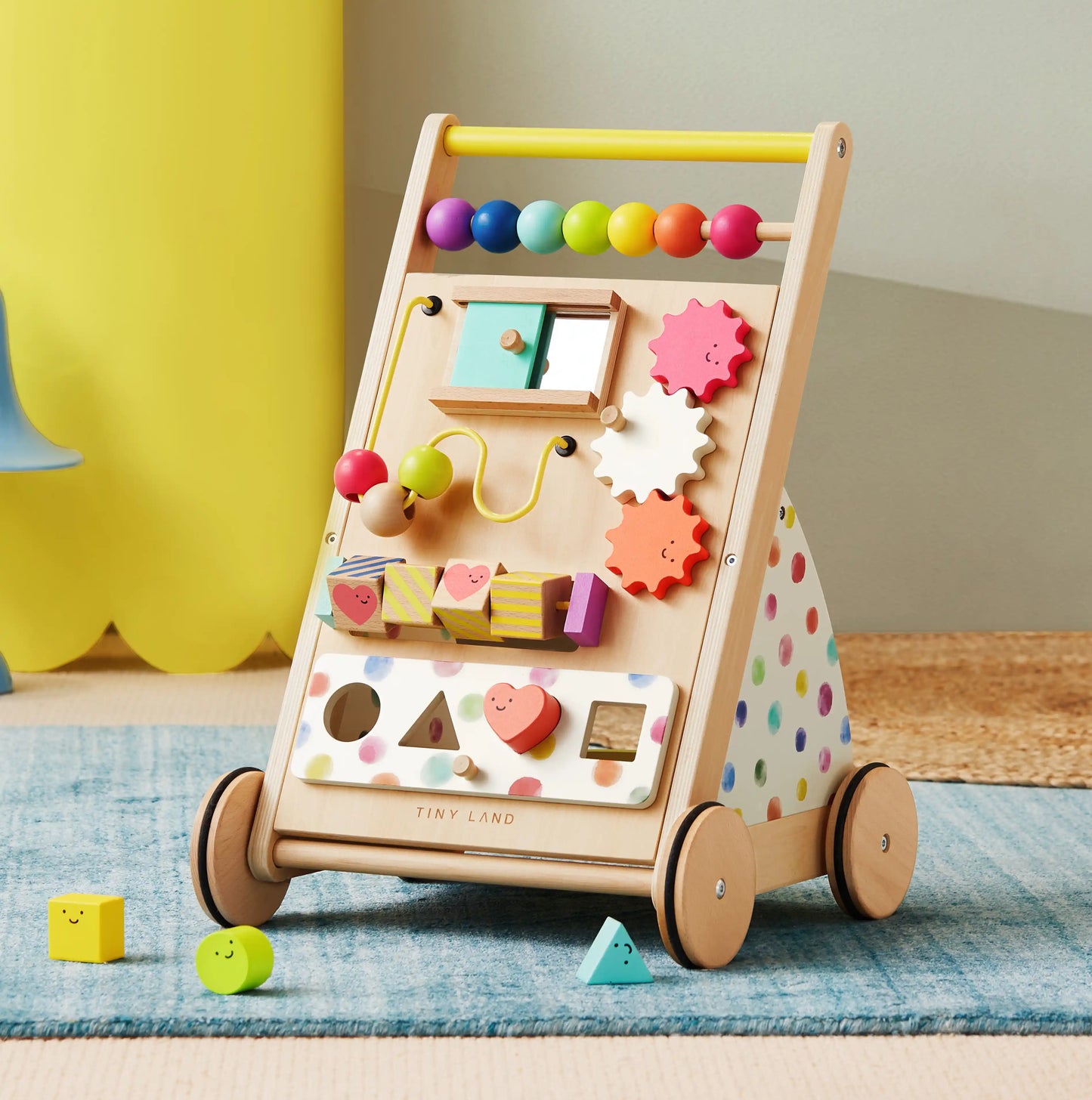 The Tiny Land® Premium Natural Wooden Activity Walker features colorful wheels, bead mazes, gears, and shape sorting blocks with polka dot sides. Various shaped blocks from the walker are scattered on the nearby floor.