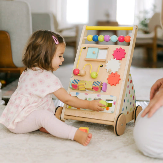 A toddler plays on the floor with the Tiny Land® Premium Natural Wooden Activity Walker, which features colorful beads, gears, and shapes. An adult sits nearby in a bright, cozy room.