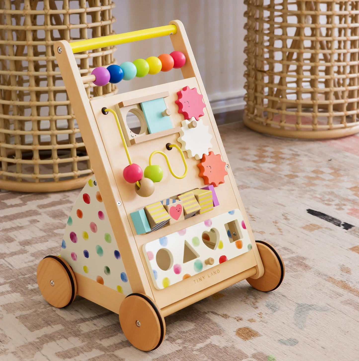 The Tiny Land® Premium Natural Wooden Activity Walker, with colorful beads, shapes, gears, a bead maze, and a shape sorter, is placed on a patterned rug in a cozy room with wicker baskets in the background.