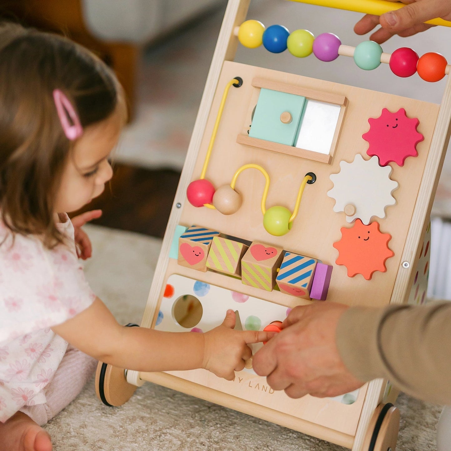 A young child plays with the Tiny Land® Premium Natural Wooden Activity Walker, featuring beads, gears, blocks, and interactive elements. An adult's hand gently guides the child's hand to a part of the wooden activity walker.