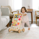A toddler in a pink outfit pushes a Tiny Land® Premium Natural Wooden Activity Walker across a bright, cozy living room filled with mid-century modern furniture and soft natural light.
