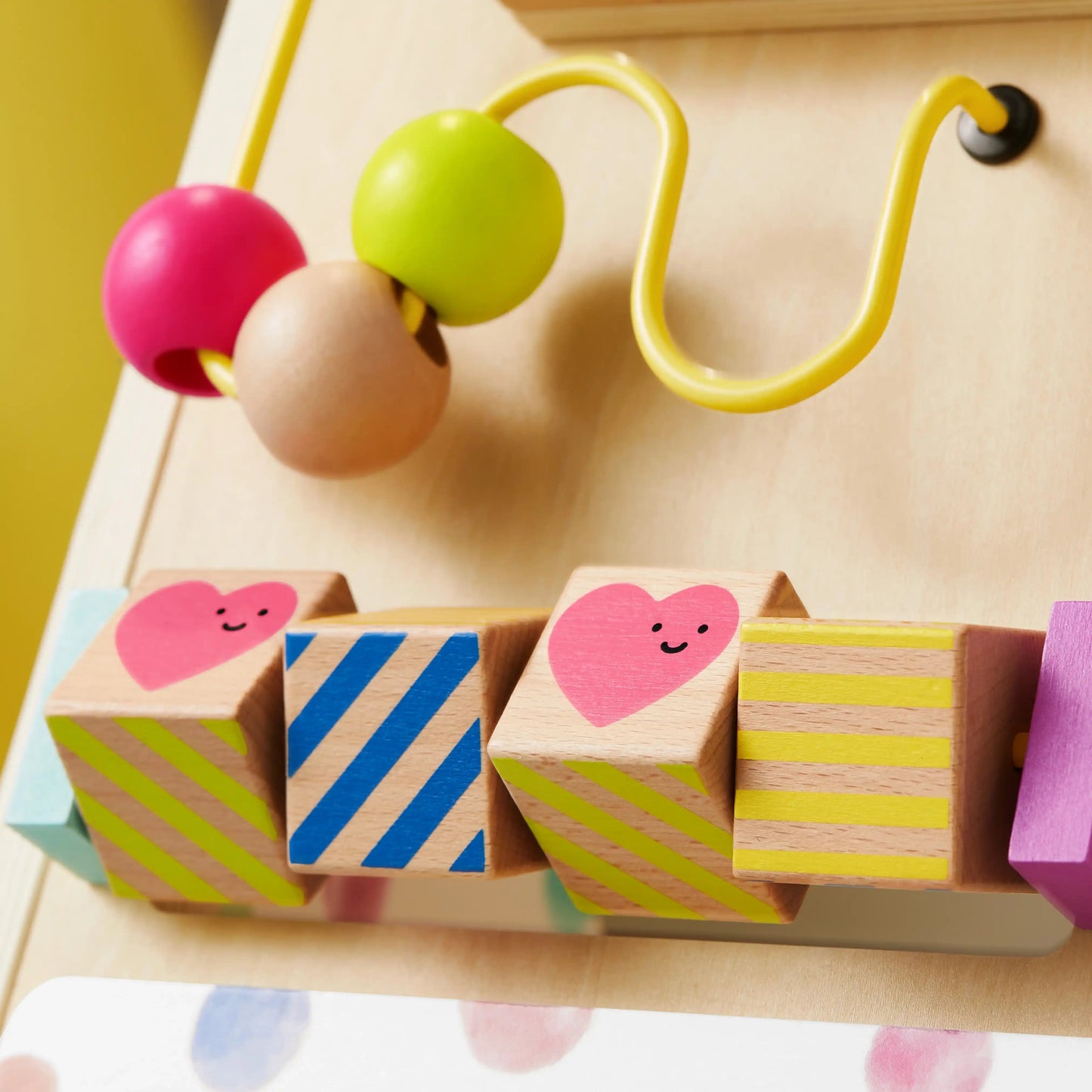 A close-up of the Tiny Land® Premium Natural Wooden Activity Walker, featuring colorful beads on a yellow wire and spinning blocks with heart and striped patterns, set against a light wood background.