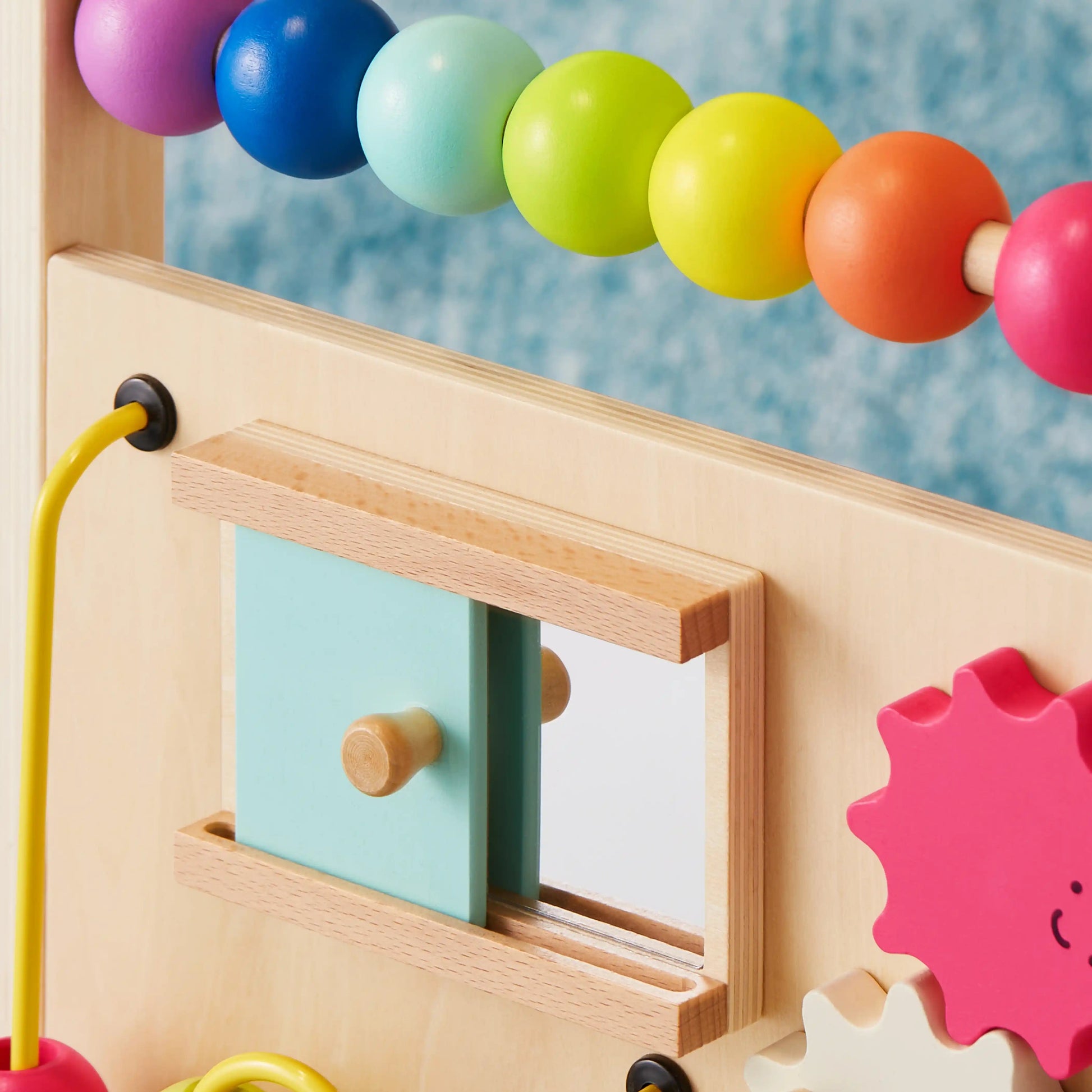 A close-up of the Tiny Land® Premium Natural Wooden Activity Walker shows colorful beads, a sliding door with a mirror, and a pink gear-shaped toy against a blurred blue background.