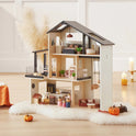 The Tiny Land® Sweetwood Edge Dollhouse, featuring multiple rooms, miniature furniture, and decor, sits on a fluffy white rug. Fall accents—like small pumpkins—and candles add a cozy touch nearby.