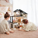 Two young girls in light dresses play on a soft carpet with the Tiny Land® Sweetwood Edge Dollhouse, arranging miniature furniture. A cozy bed and pastel decor set a charming scene for their creative play.