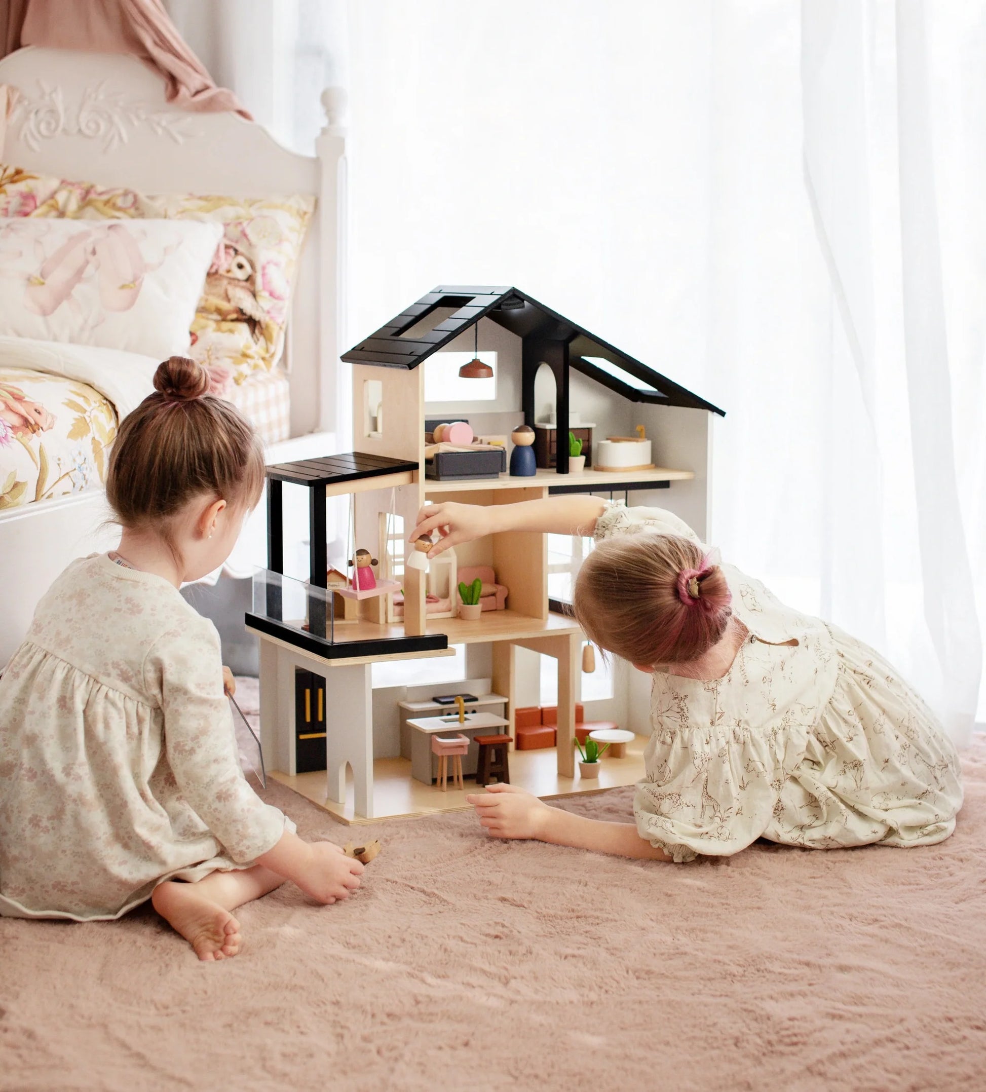 Two young girls in light dresses play on a soft carpet with the Tiny Land® Sweetwood Edge Dollhouse, arranging miniature furniture. A cozy bed and pastel decor set a charming scene for their creative play.