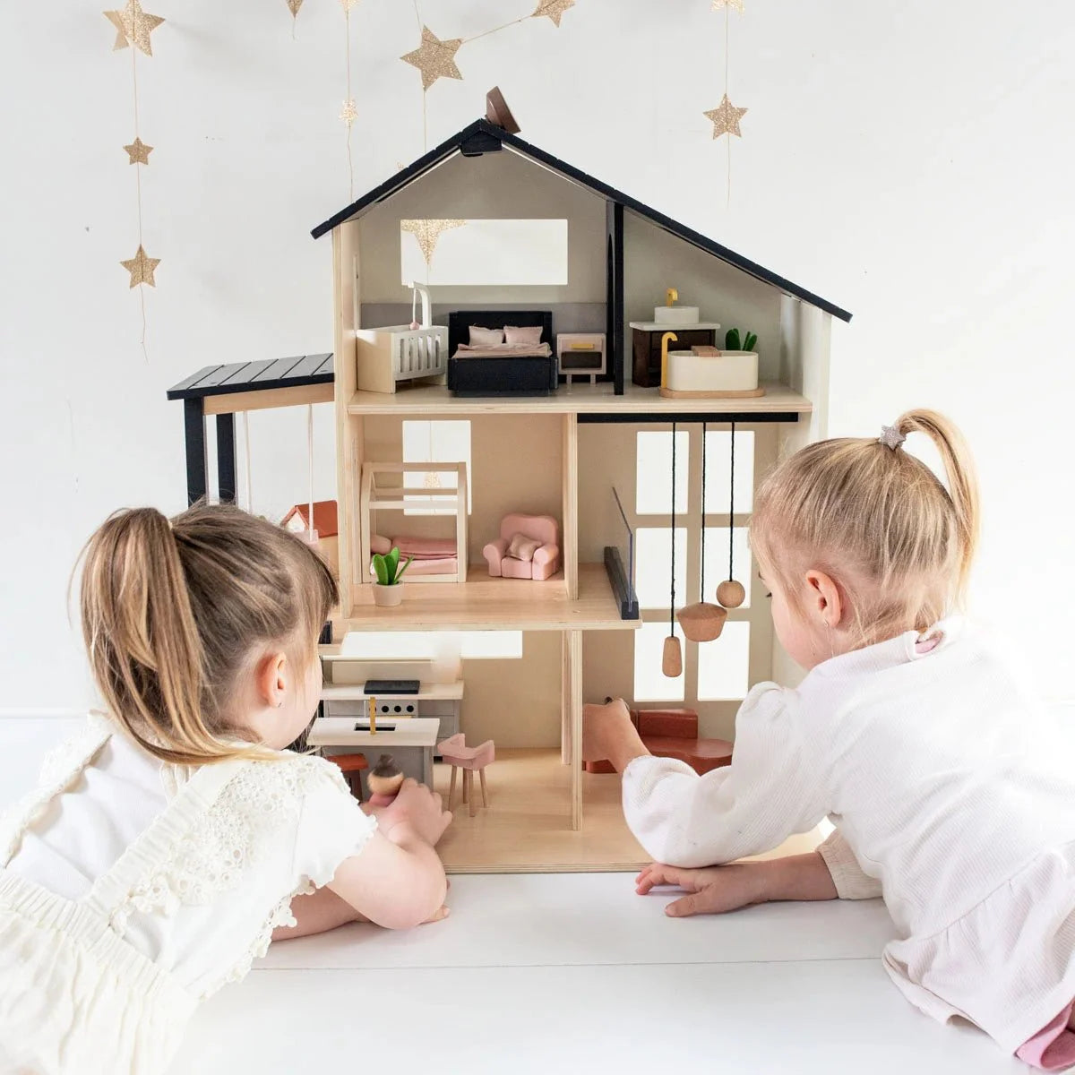 Two young children with blonde hair play with the Tiny Land® Sweetwood Edge Dollhouse, arranging furniture in its detailed multi-room interior. Gold star decorations can be seen hanging in the background.
