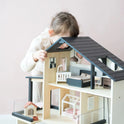 A young child leans over and peers inside the Tiny Land® Sweetwood Edge Dollhouse, closely examining the miniature furniture and rooms. The background is a plain, light pink wall.