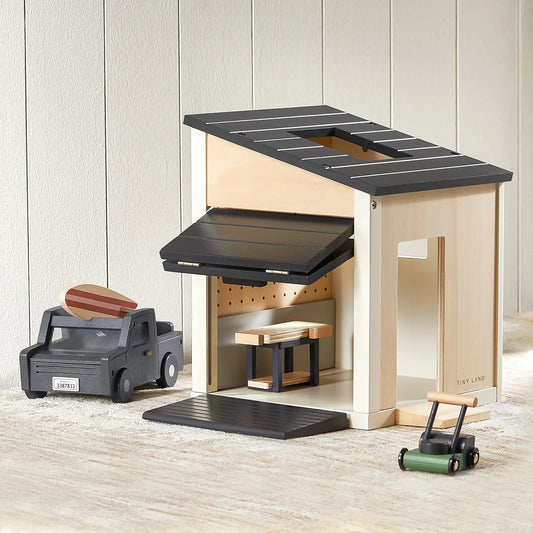 A Wooden Dollhouse Garage with Truck featuring a roll-up door, a black toy truck with a surfboard on top, and a small green push toy parked outside.