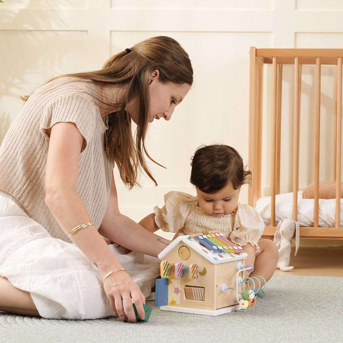 A woman and a young child sit on the floor playing with a Montessori Cottage Activity Cube – Morandi.