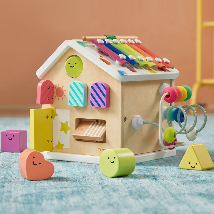 Montessori Cottage Activity Cube – Rainbow: a wooden house-shaped activity cube featuring a xylophone roof, bead maze, and opening doors, surrounded by small geometric blocks with smiley faces designed to develop fine motor skills.