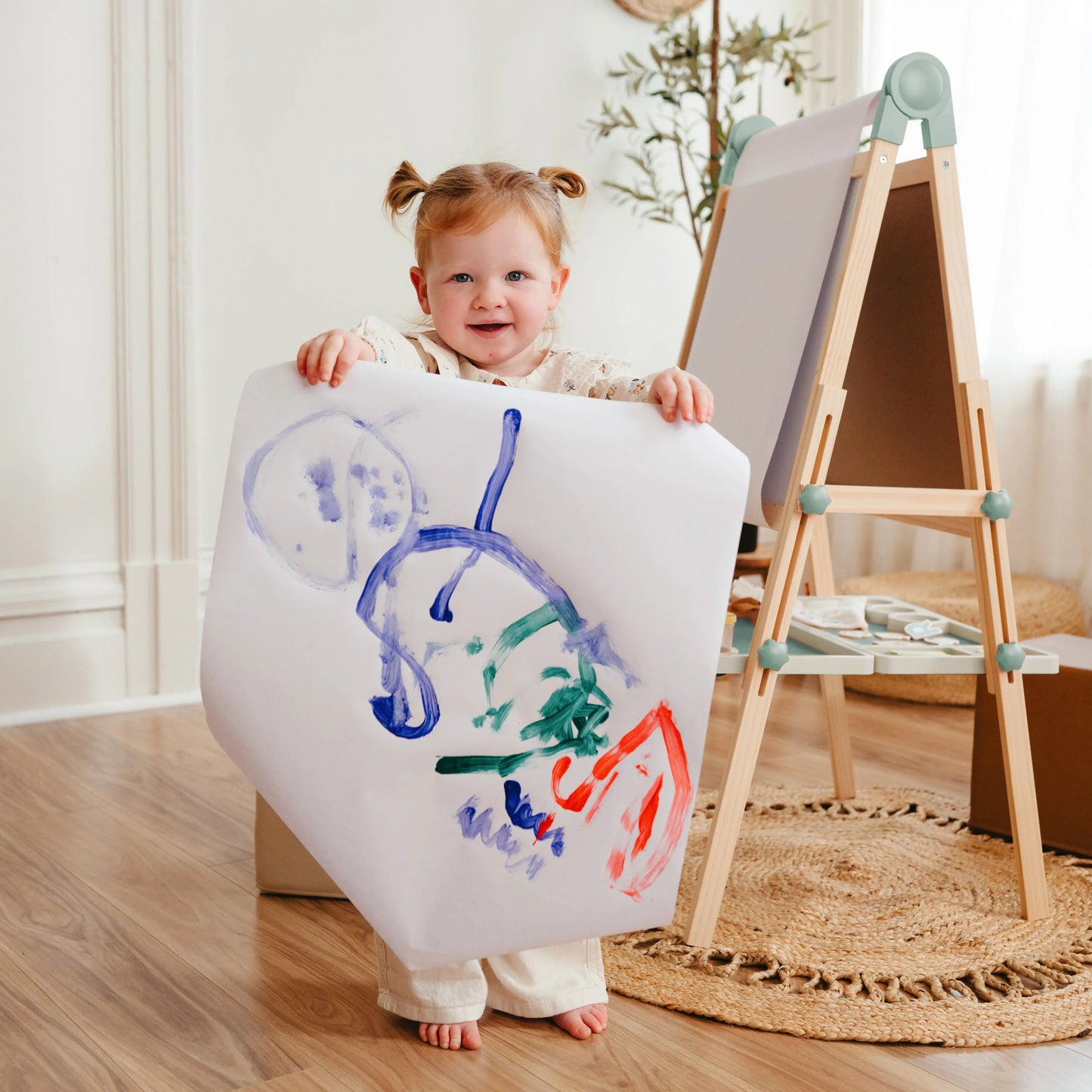 A young child with pigtails smiles indoors, holding a colorful abstract art sheet while standing in front of the Tiny Land® Magnitales Easel for Kids - Green in a cozy, well-lit room.