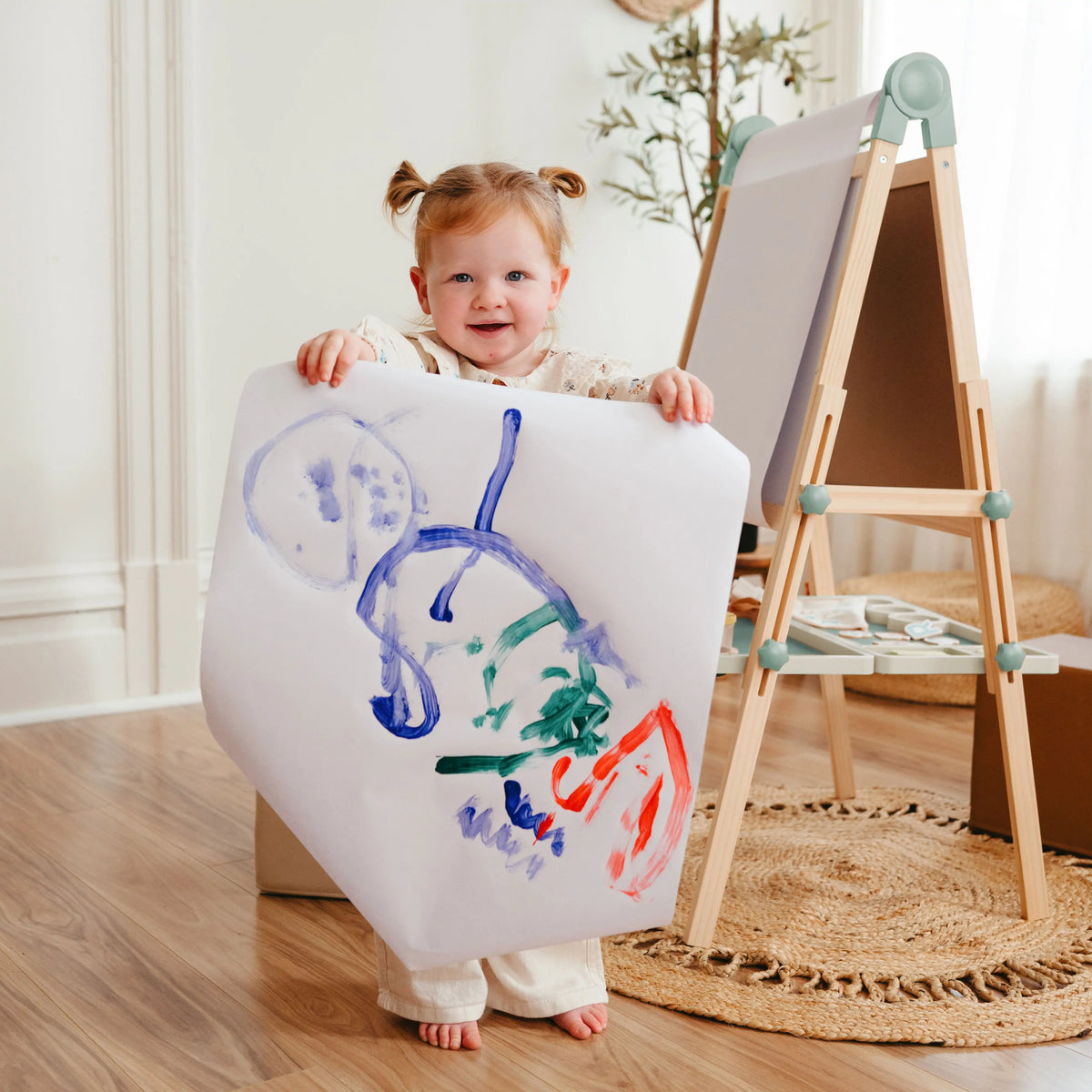 A toddler stands in front of a Magnitales Double-Sided Art Easel with Paper Roll, holding up a large sheet of paper with colorful finger painting.