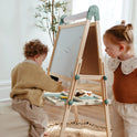 Two young children use the Tiny Land® Magnitales Easel for Kids - Green to draw and play with art supplies at its adjustable whiteboard in a cozy, sunlit room with a plant in the background.