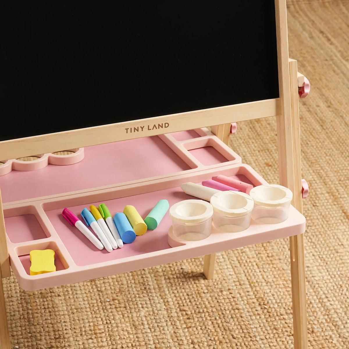 The Magnitales Double-Sided Art Easel with Paper Roll features a wooden frame, a black chalkboard, and a pink storage tray holding markers, chalk, a yellow eraser, and three empty paint cups for kids.