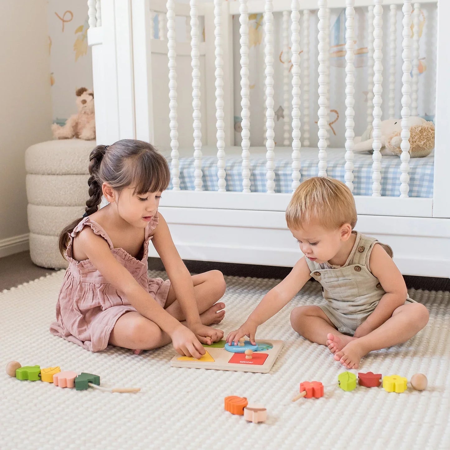 Two young children play on a soft rug by a white crib, exploring Tiny Land® Montessori Toys for Toddlers (18–24 Months) – Montessori Toys for 2 Year Olds. The girl points at the puzzle as the boy watches attentively.
