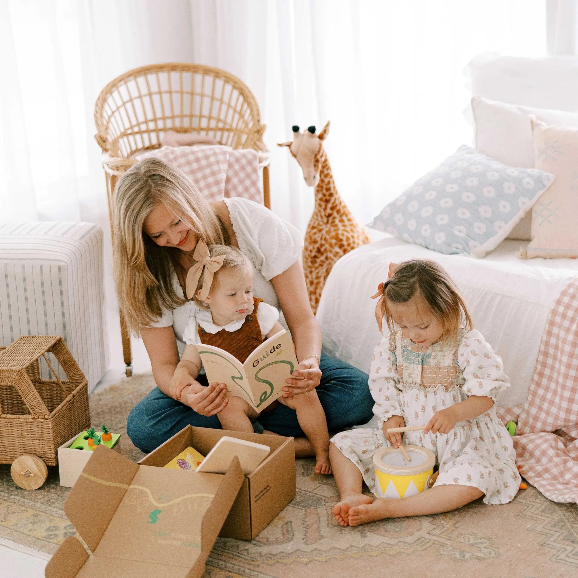 A woman sits on the floor with two young children in a bright, cozy room. One child (13–15 months) sits on her lap holding a book, while the other plays with the Tiny Land® Montessori Toys for Toddlers. A toy giraffe and boxes are nearby.