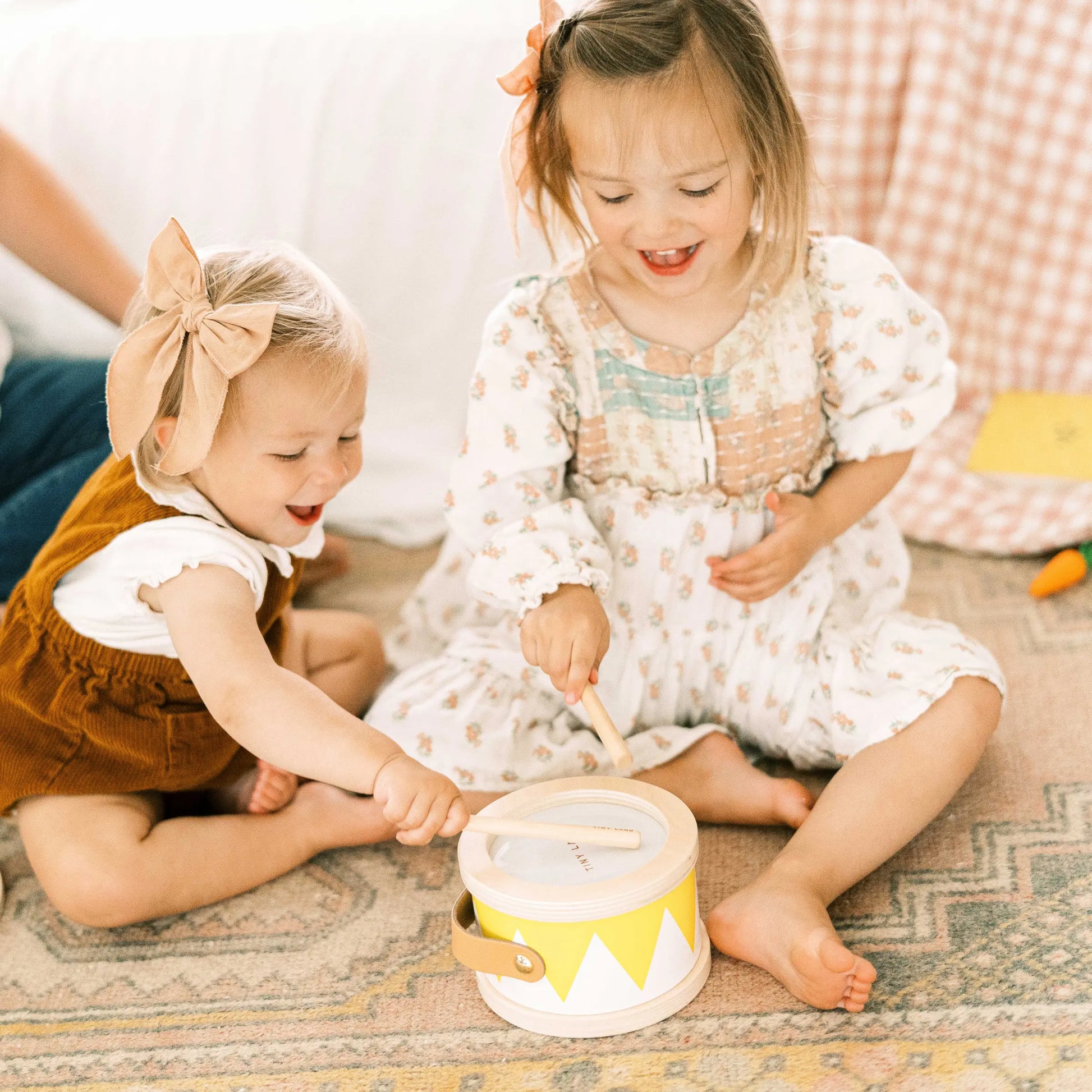 Two young girls, 13–15 months old, sit on a rug smiling and playing with a drum from the Tiny Land® Montessori Toys for Toddlers. Both wear light-patterned outfits with bows, happily engaging with this educational toy.