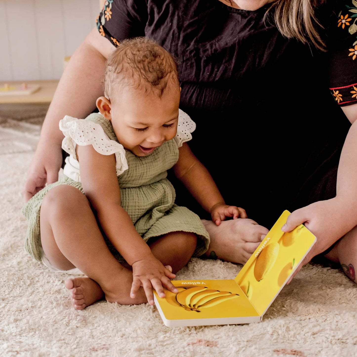 A smiling 13–15-month-old baby sits on a carpet, exploring a picture book with a banana illustration, as an adult in a black dress with embroidered sleeves sits beside them. The Tiny Land® Montessori Toys for Toddlers box is nearby.