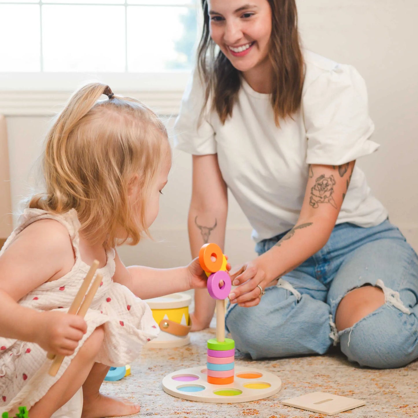 A young woman smiles as she plays with a 13–15 month old toddler, who is stacking colorful wooden rings from the Tiny Land® Montessori Toys for Toddlers in a bright room.