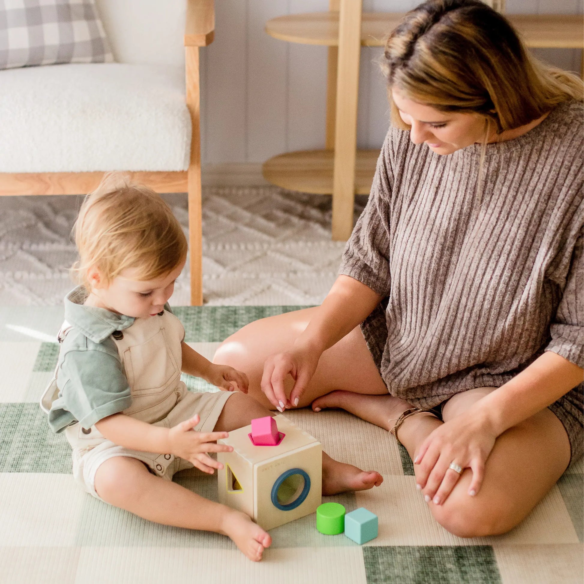 A toddler and an adult sit on a checkered mat, playing with the Tiny Land® Montessori Toys for Toddlers (16–18 Months), fitting colorful shapes into matching holes—an ideal educational toy for early learning.