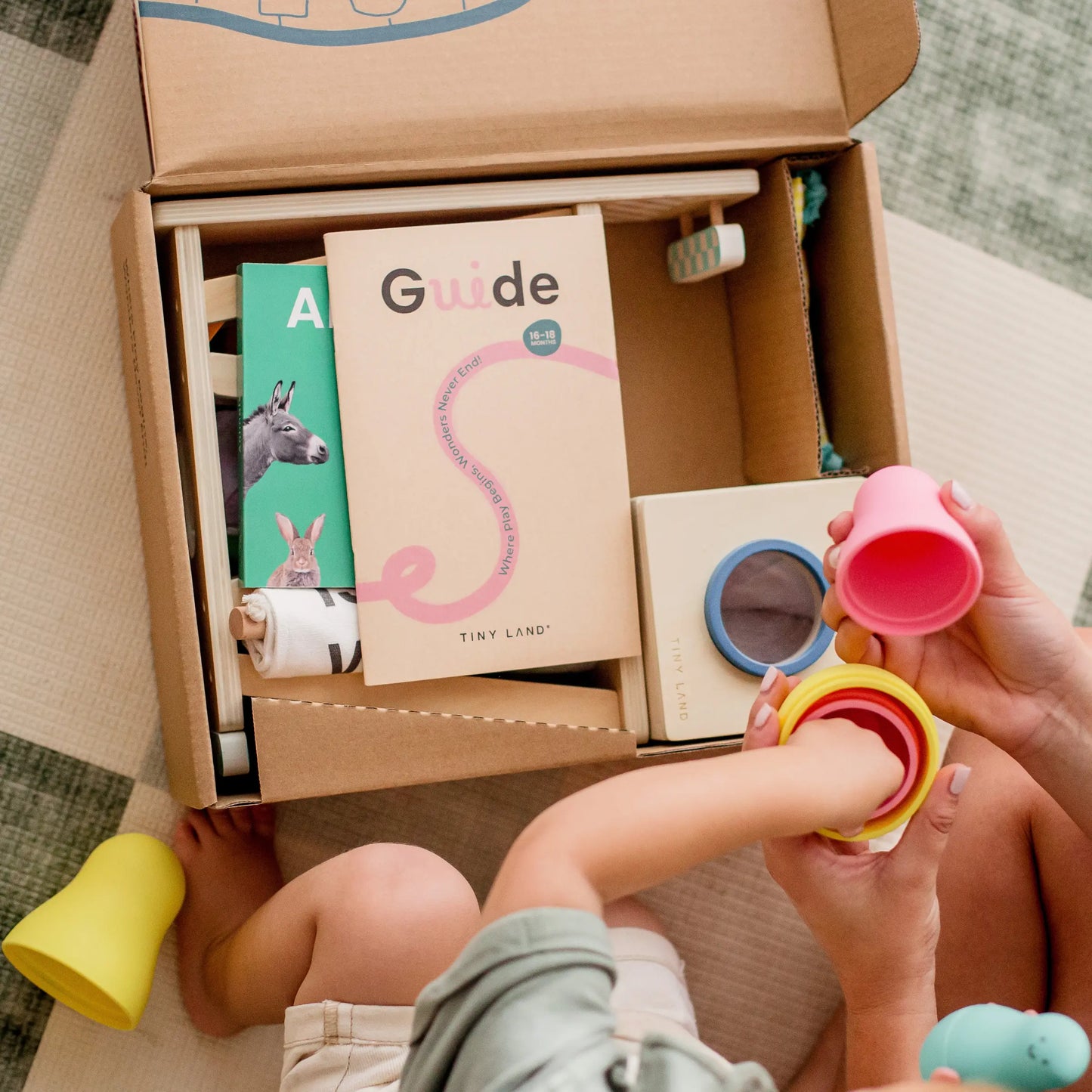 A child and adult play with colorful stacking cups beside the open Tiny Land® Montessori Toys for Toddlers (16–18 Months) box, featuring a guide booklet, a green animal book, and educational toys on a checkered floor.