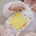 A baby in white holds a soft, polka-dotted cloth with a yellow patch while lying beside Tiny Land® Montessori Toys for Infants (10–12 Months) on a light textured surface.