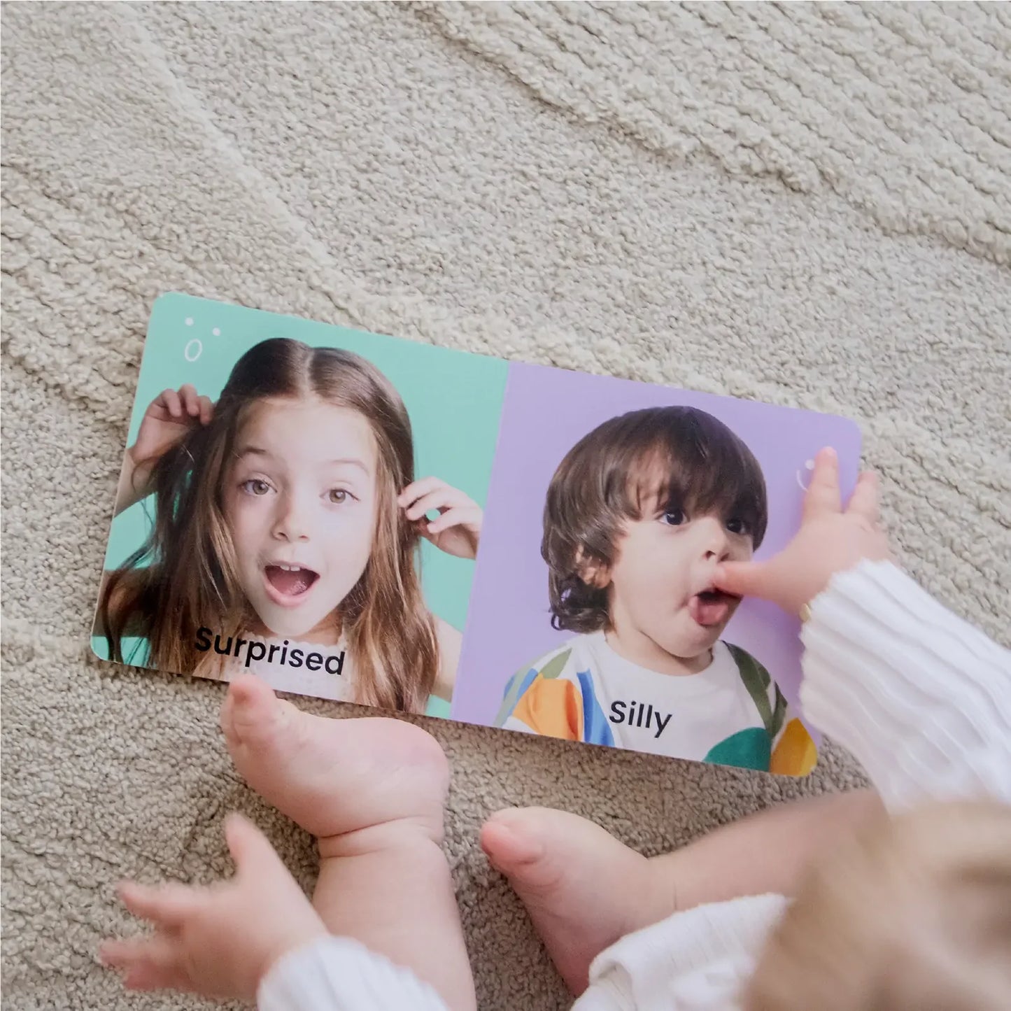 A baby sits on a carpet exploring a board book page with photos labeled Surprised and Silly, while Tiny Land® Montessori Toys for Infants (10–12 Months) nearby create an ideal scene of learning and discovery.