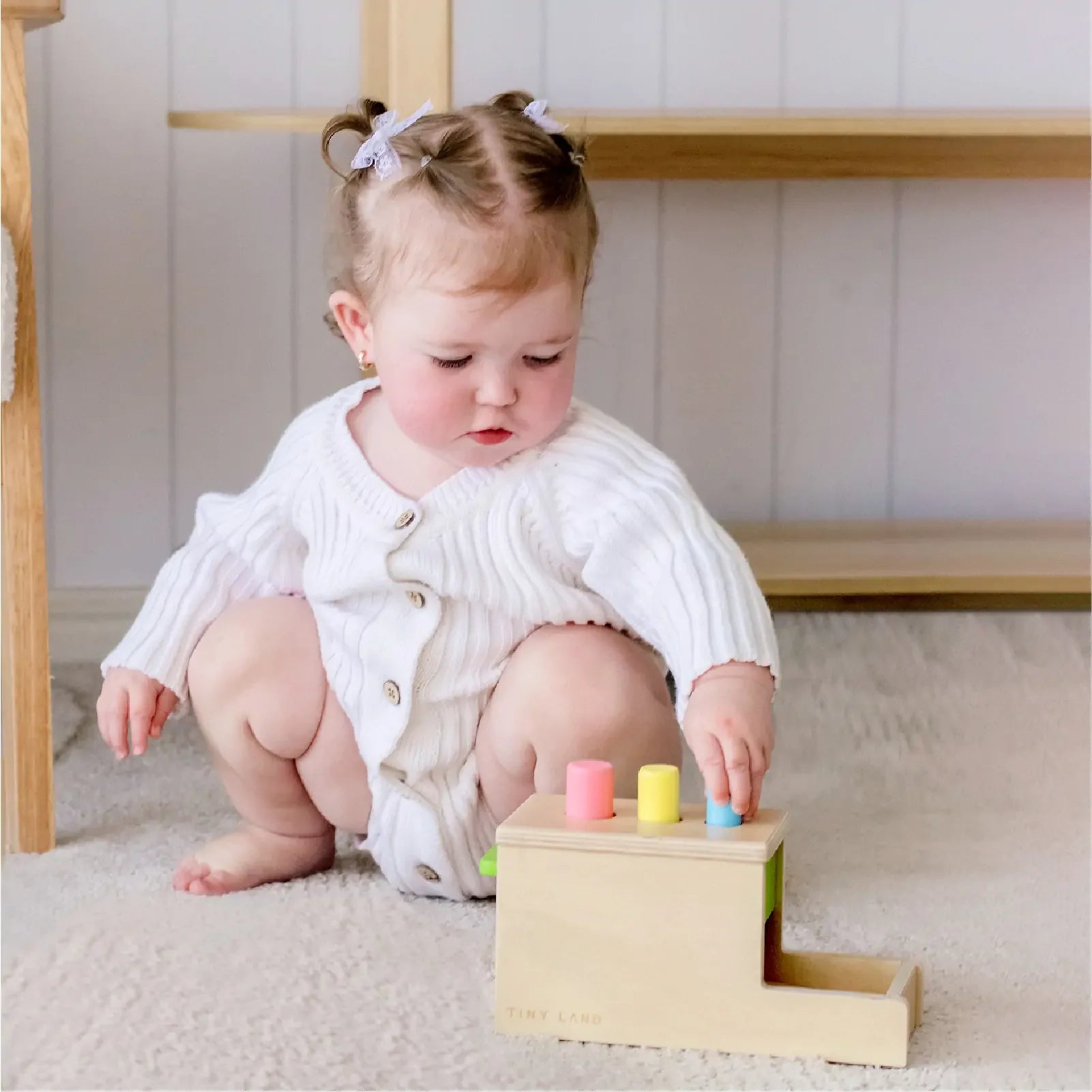 A baby in a white sweater and bow hair clips sits on a soft carpet, playing with the Tiny Land® Montessori Toys for Infants (10–12 Months), a colorful wooden playset designed for baby development.
