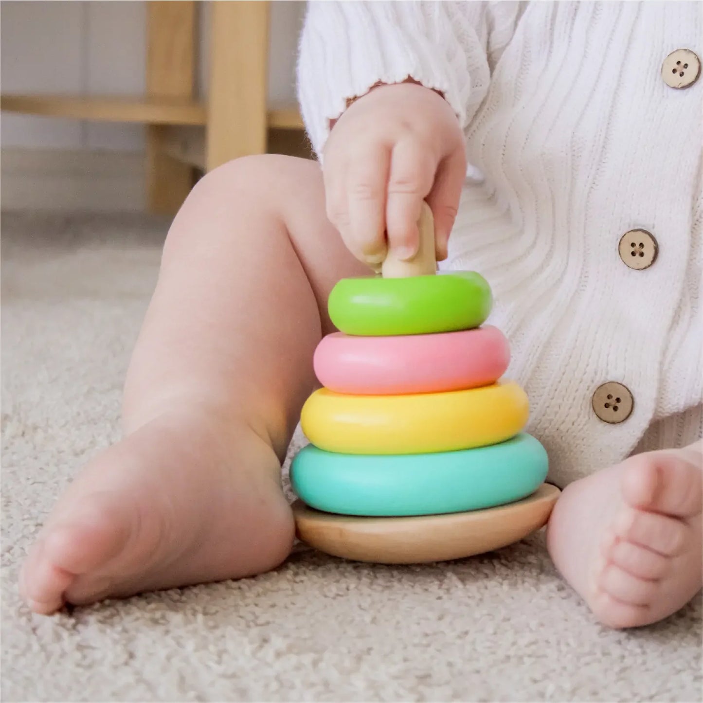 A baby in a white sweater plays on a carpet with Tiny Land® Montessori Toys for Infants (10–12 Months), stacking four colorful wooden rings—green, pink, yellow, and blue—onto a sturdy wooden base.