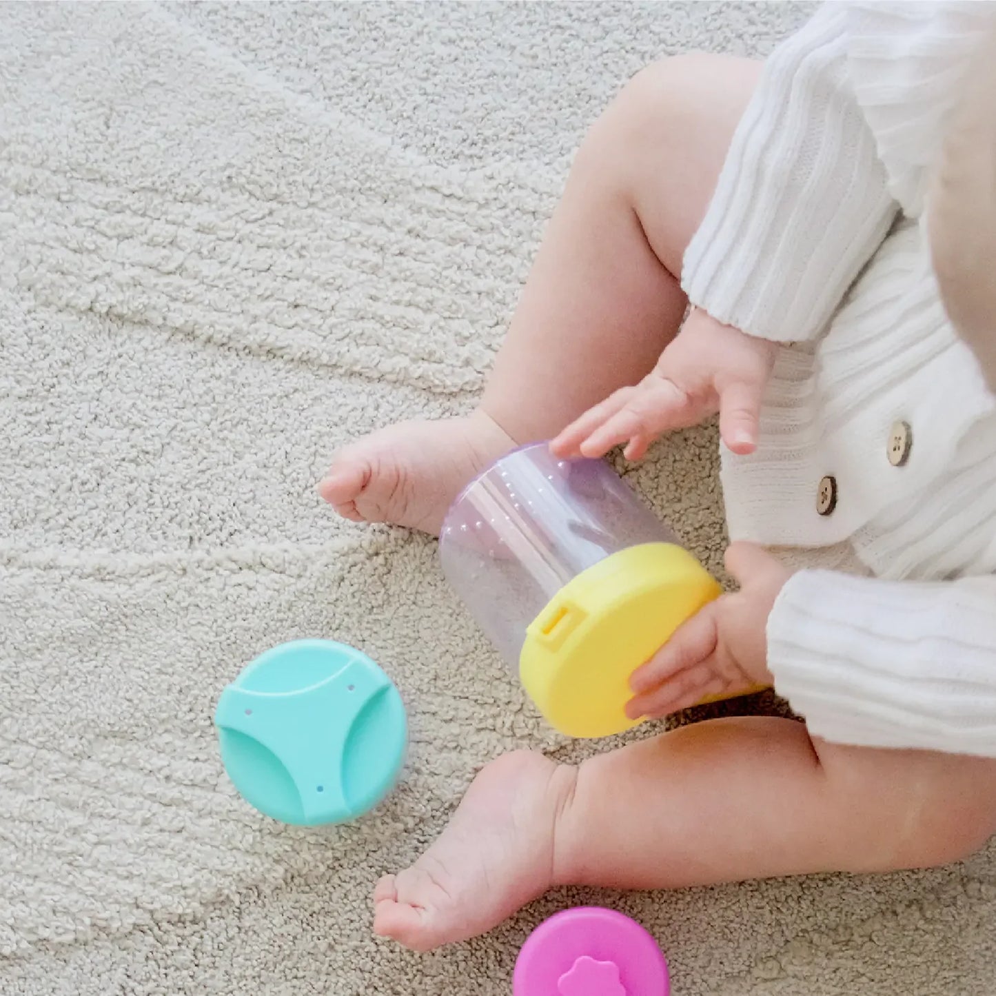 A baby’s lower body and hands are shown as they play on a beige carpet with Tiny Land® Montessori Toys for Infants (10–12 Months), exploring a clear container with a yellow lid and colorful lids nearby to encourage hands-on learning.