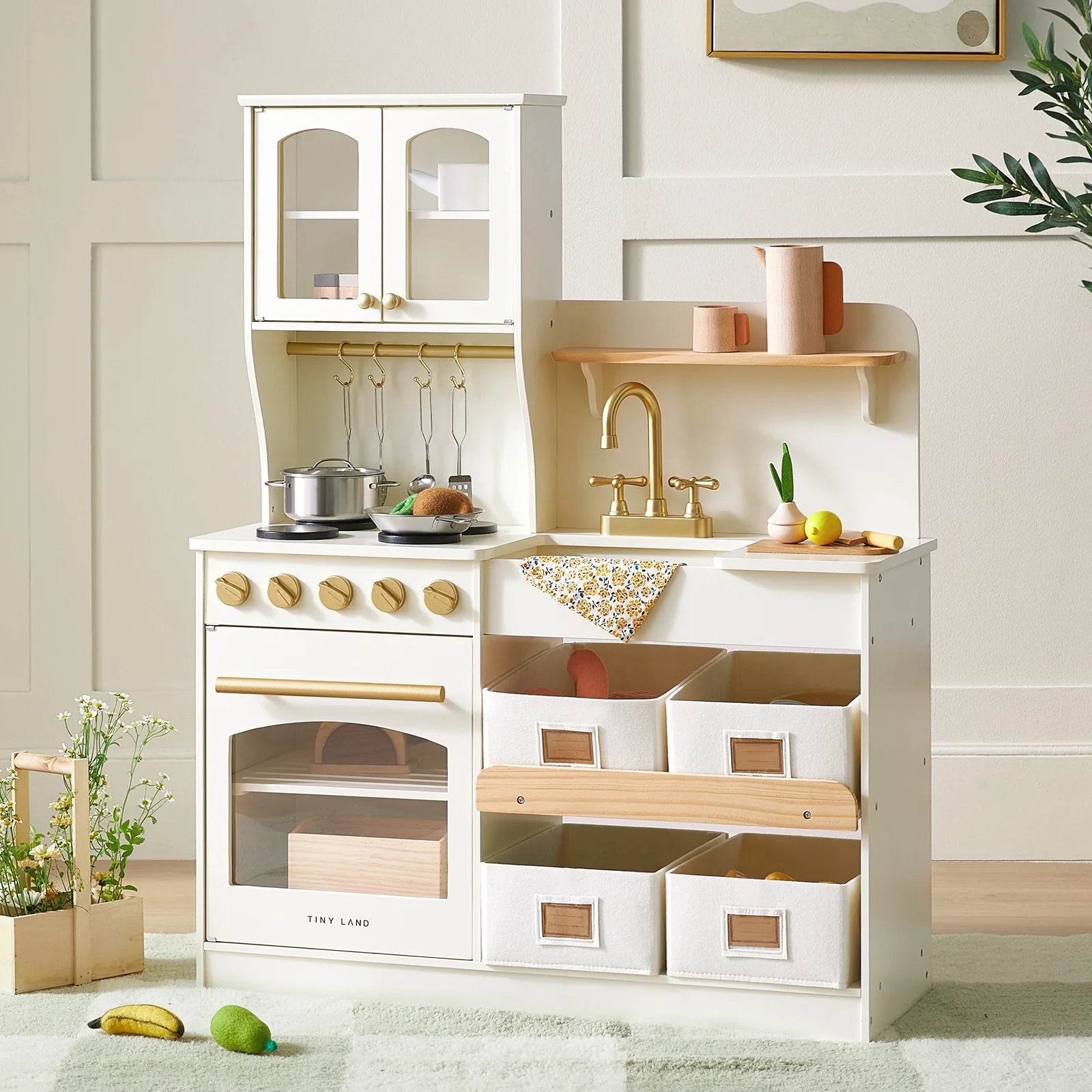 The Tiny Land® Wooden Play Kitchen with Storage for Creative Toddlers features a stove, oven, sink with gold faucet, hanging cookware, pot, plant, and six labeled fabric bins—perfect for light rooms with minimal decor.