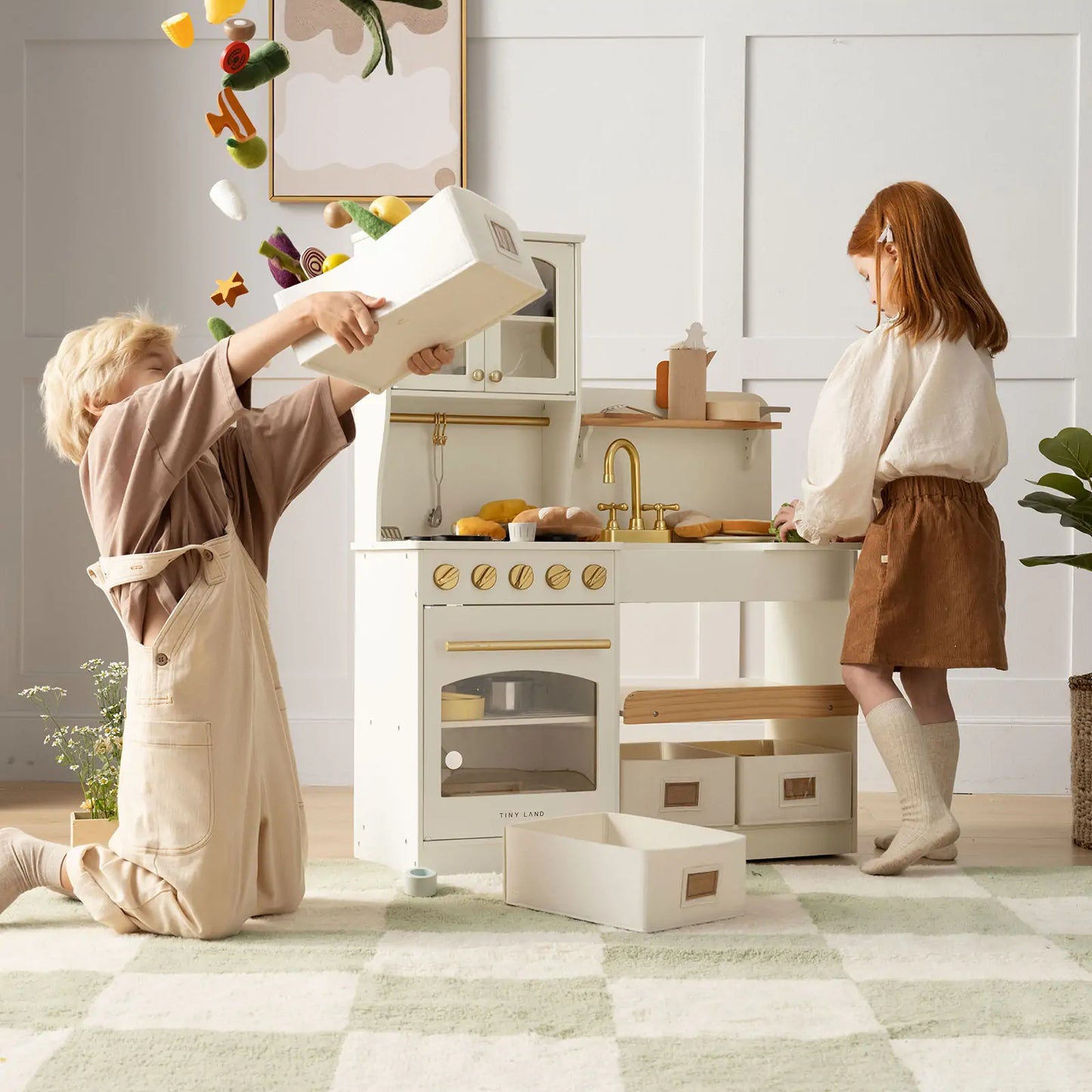 Two children enjoy the Tiny Land® Wooden Play Kitchen with Storage for Creative Toddlers; one tosses toy food while the other watches, both playing in a bright, cozy room filled with pretend cookware.