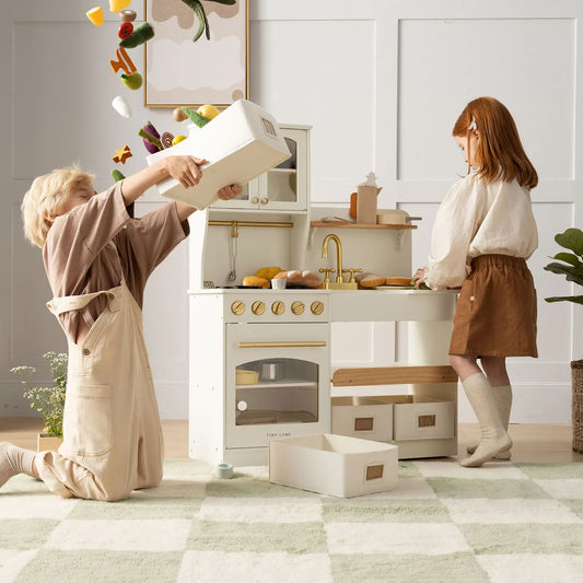 Two children enjoy the Tiny Land® Wooden Play Kitchen with Storage for Creative Toddlers; one tosses toy food while the other watches, both playing in a bright, cozy room filled with pretend cookware.