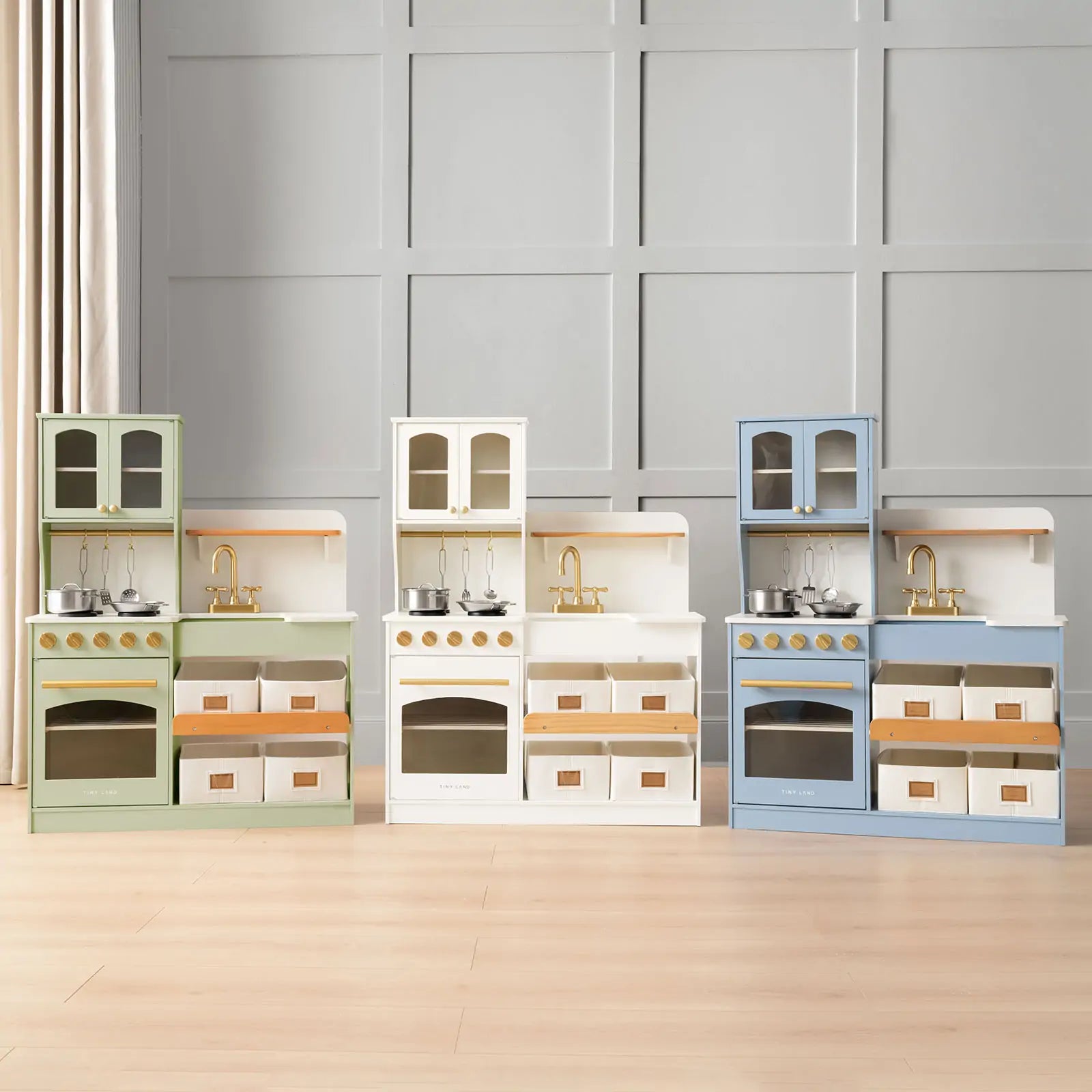 Three Tiny Land® Wooden Play Kitchens with storage in pastel green, white, and blue stand side by side on a light wood floor, featuring stove, oven, sink, and gold-tone knobs and faucet for creative toddler play.