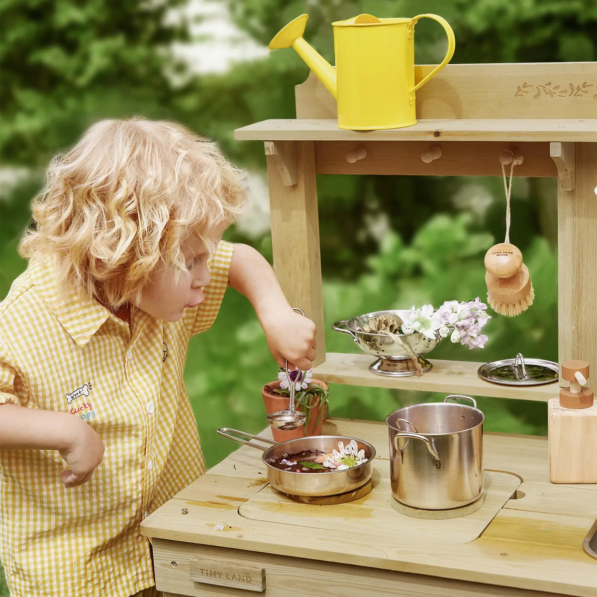 A child engages in outdoor sensory play at a DuoPlay Outdoor Mud Kitchen with Working Faucet, stirring a metal pot filled with water and flowers.
