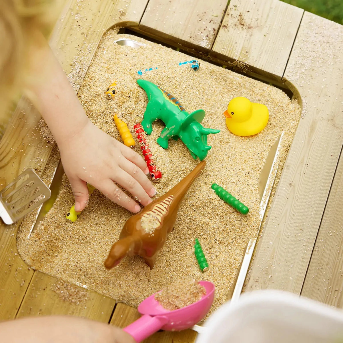 A child’s hand plays with toy dinosaurs, a rubber duck, and small plastic figures in the metal tray of a DuoPlay Outdoor Mud Kitchen with Working Faucet, filled with sand for sensory play.