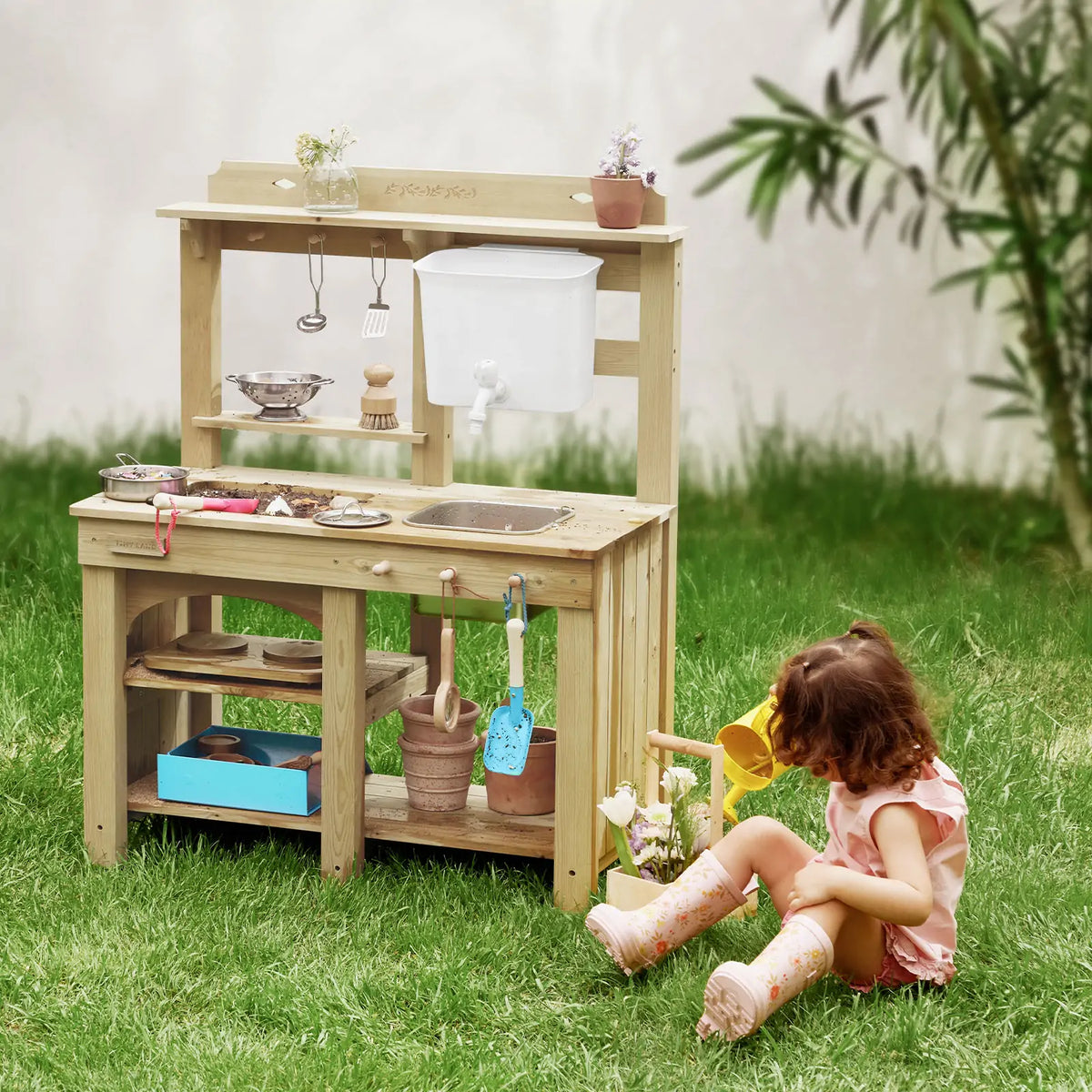 A child sits on the grass engaging in sensory play with a DuoPlay Outdoor Mud Kitchen with Working Faucet featuring a basin, water reservoir, tap, shelving, and assorted plastic kitchen utensils and pots.