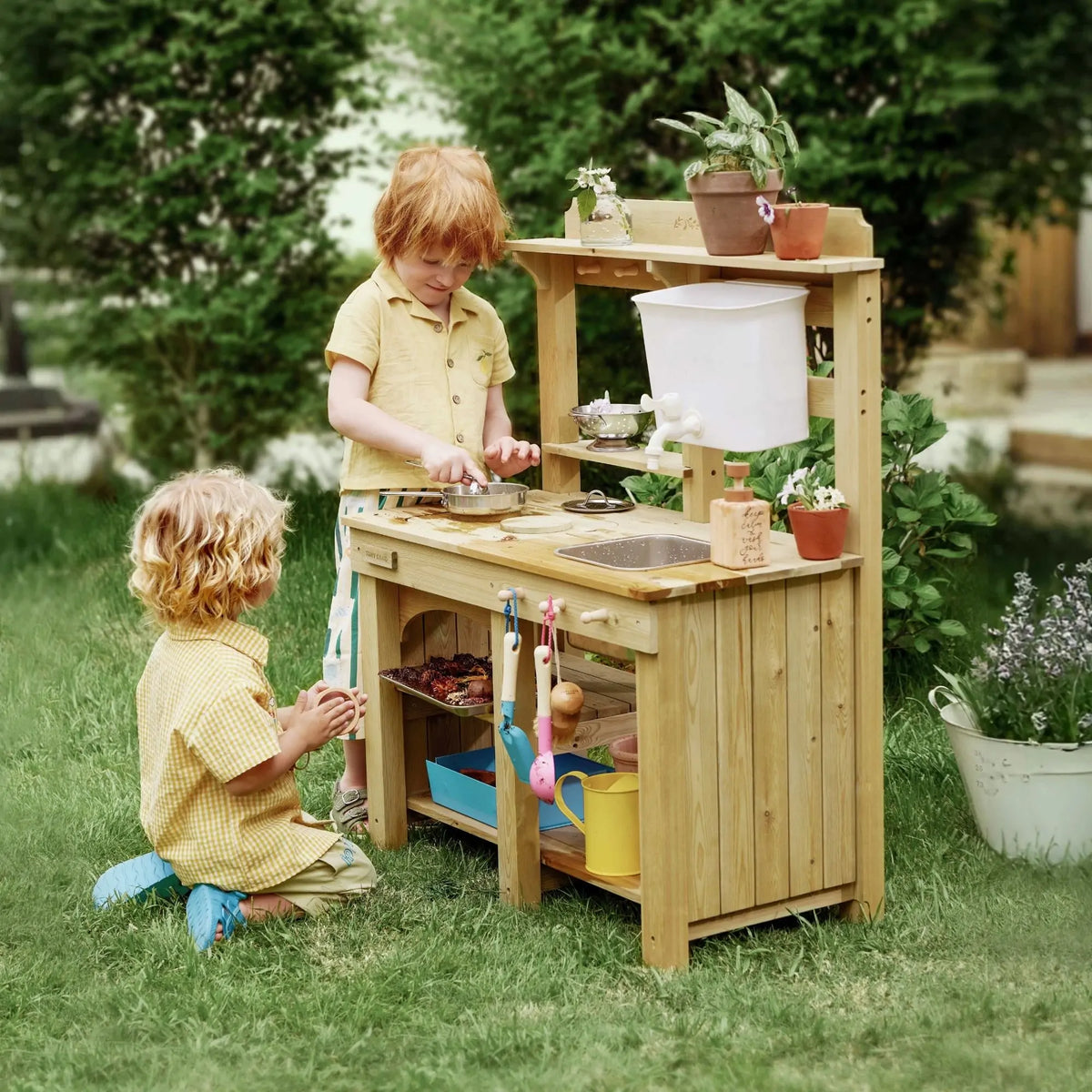 Two children engage in outdoor sensory play with a DuoPlay Outdoor Mud Kitchen with Working Faucet, which features a wooden design, sink, faucet, and cooking utensils in a grassy yard.