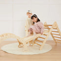 A girl sits on the wooden slide of a 7-in-1 Montessori Climbing Set with Play Pad, while a boy stands beside her.