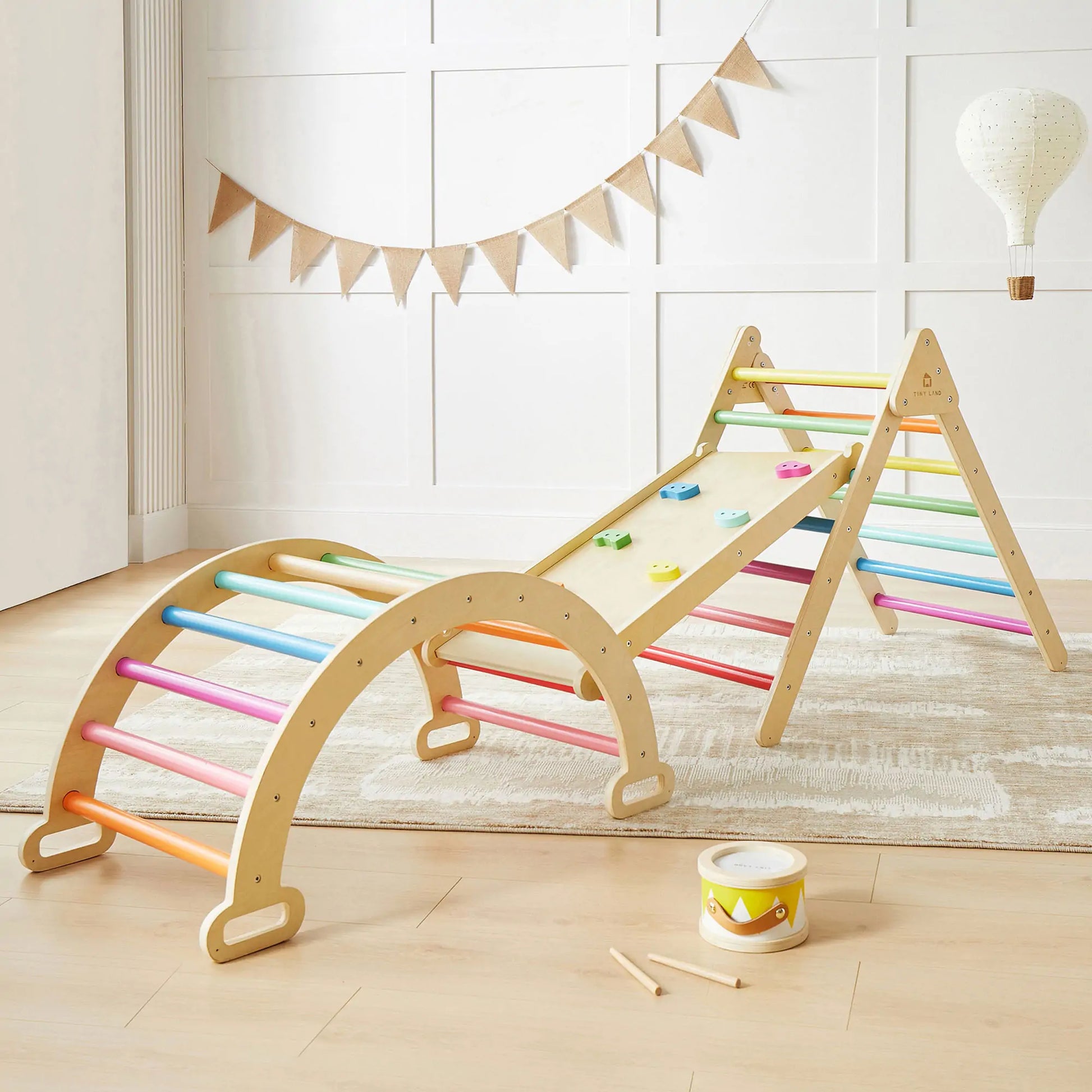 The Tiny Land® 7-in-1 Rainbow Climbing Set, featuring a Pikler Triangle, climbing arch, and a ramp with colorful holds, sits on a light rug in a bright room with white walls and playful decor.