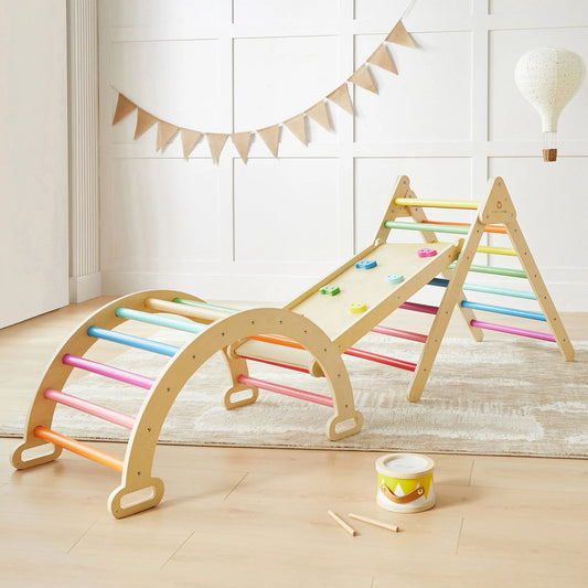 The Tiny Land® 7-in-1 Rainbow Climbing Set, featuring a Pikler Triangle, climbing arch, and a ramp with colorful holds, sits on a light rug in a bright room with white walls and playful decor.