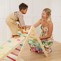 A young child climbs the Tiny Land® 7-in-1 Rainbow Climbing Set as a woman in a floral dress kneels nearby, smiling and supporting the child in a bright, cozy room.