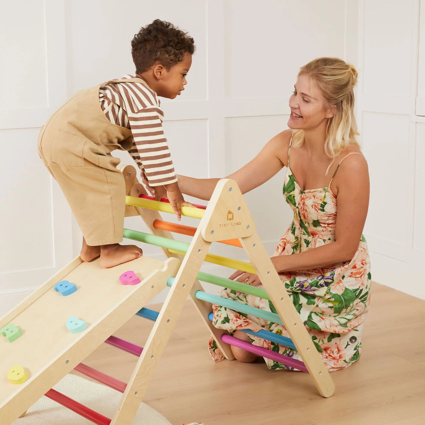 A young child climbs the Tiny Land® 7-in-1 Rainbow Climbing Set as a woman in a floral dress kneels nearby, smiling and supporting the child in a bright, cozy room.
