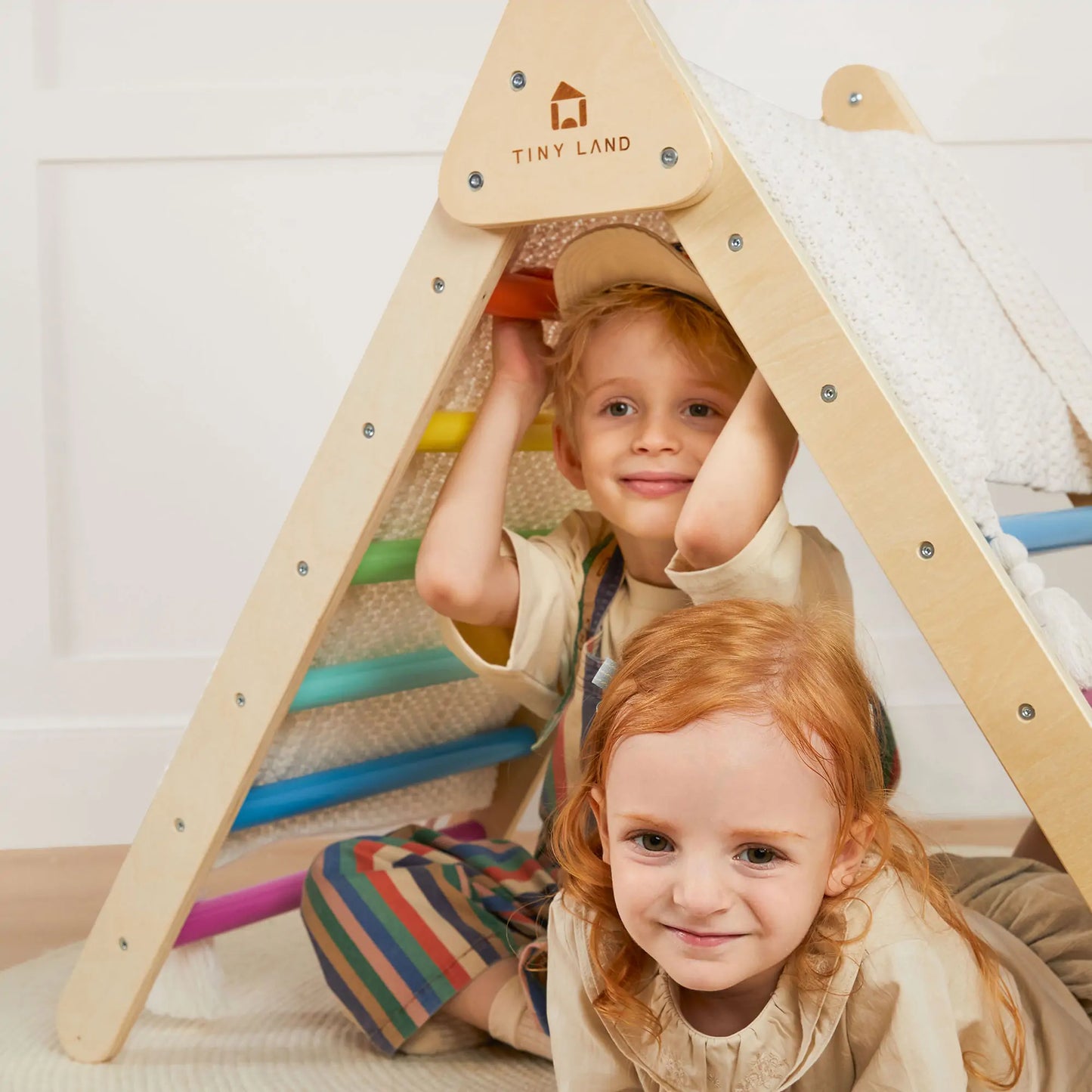 Two young children smile and play with a Tiny Land® 7-in-1 Rainbow Climbing Set. One sits inside the colorful Pikler Triangle while the other crouches outside, enjoying endless indoor fun with this vibrant climbing toy.
