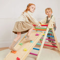 Two young children play in a bright indoor room on the Tiny Land® 7-in-1 Rainbow Climbing Set, featuring a wooden Pikler Triangle with colorful rungs. The girl climbs up while the boy holds onto the sturdy structure.