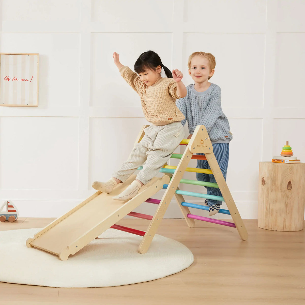 A child slides down the wooden ramp of a 7-in-1 Montessori Climbing Set with Play Pad – Rainbow, which features colorful rungs, while another child stands behind them on the padded play cushion.