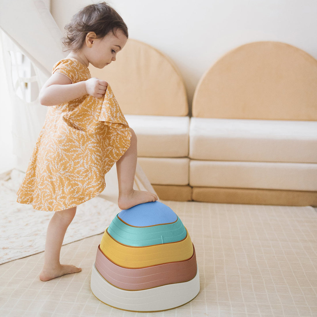 A toddler in a yellow floral dress steps onto a stack of four nested, bowl-shaped Stacking Balance Stepping Stones – Beach in shades of blue, yellow, terracotta, and cream.