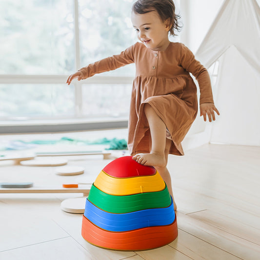 A young child balances on Stacking Balance Stepping Stones – Rainbow, a stack of colorful, bowl-shaped stepping stones, practicing their balance and coordination.